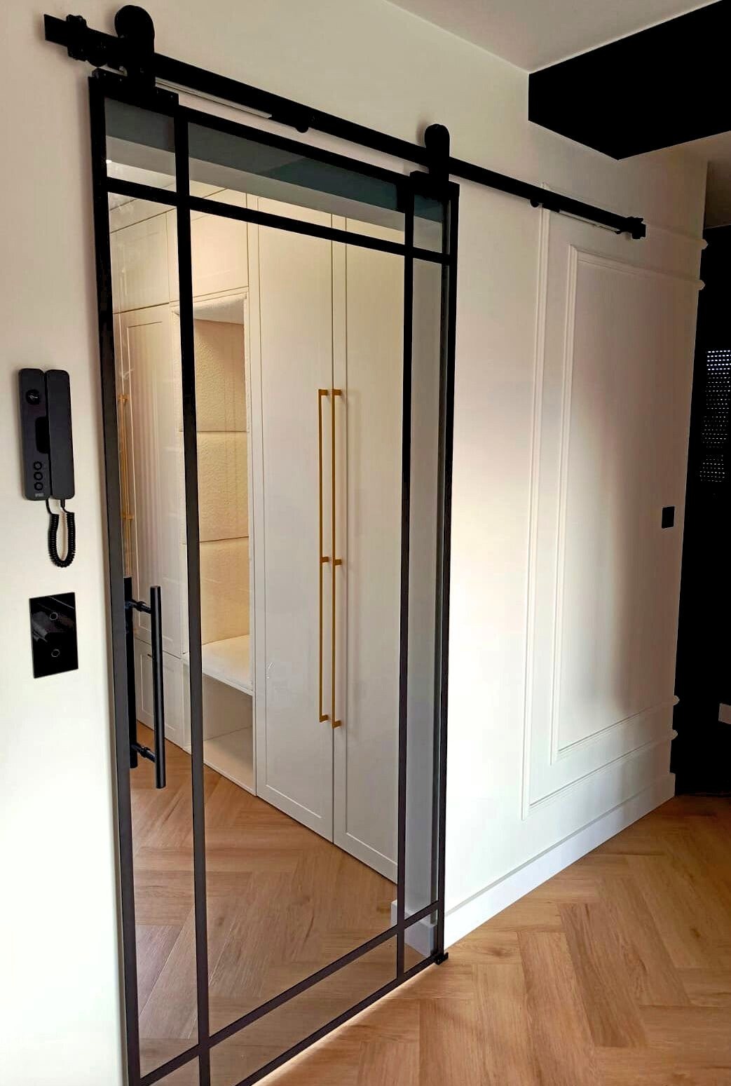 Przesuwne drzwi szklane z czarną ramą w Wieruszowie. Widoczny parkiet ułożony w jodełkę, białe ściany z dekoracyjnymi listwami i szafa wnękowa z złotymi uchwytami.