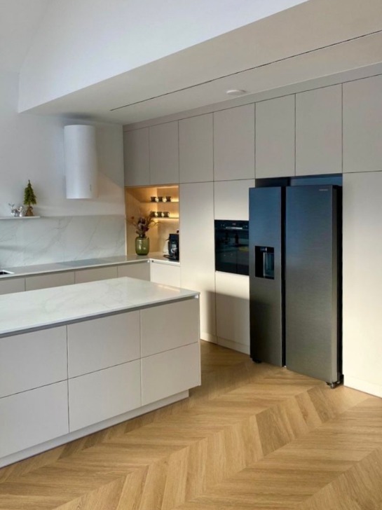 Jasna kuchnia z wyspą i parkietem w jodełkę. Minimalistyczne szafki, nowoczesny okap cylindryczny i duża lodówka side-by-side. Dekoracje i oświetlenie punktowe.