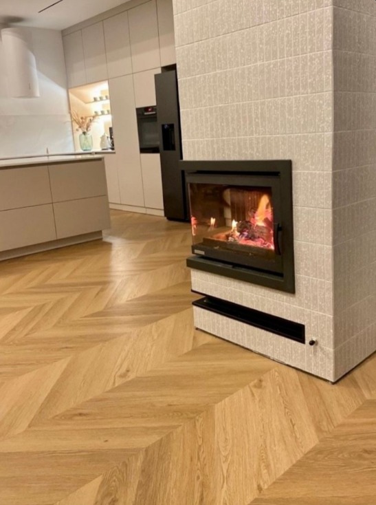 Jasny parkiet ułożony w jodełkę francuską, uzupełniony o nowoczesny kominek z obudową z płytek. W tle widoczna zabudowa kuchenna w neutralnych barwach.
