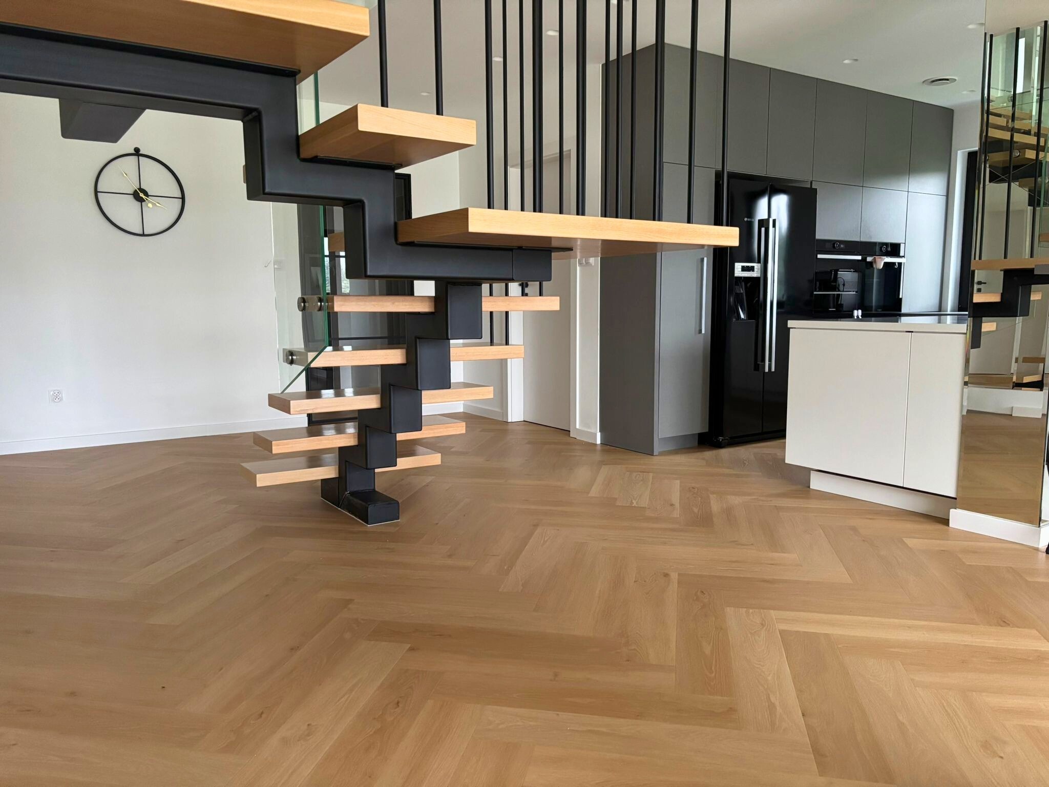 Parkiet ułożony w jodełkę francuską, widoczne schody z drewnianymi stopniami i czarną metalową konstrukcją. W tle nowoczesna kuchnia w szaro-czarnej kolorystyce.
