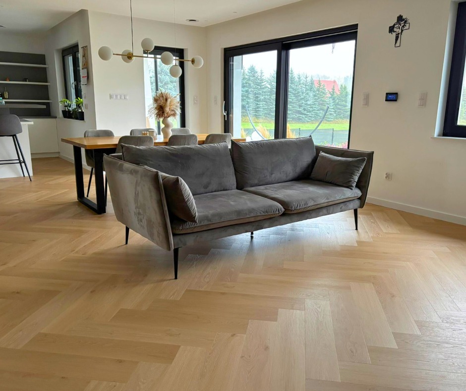 Jasny parkiet ułożony w jodełkę francuską w nowoczesnym salonie z szarą sofą i dużymi oknami. Widok na zewnątrz z zielenią drzew.