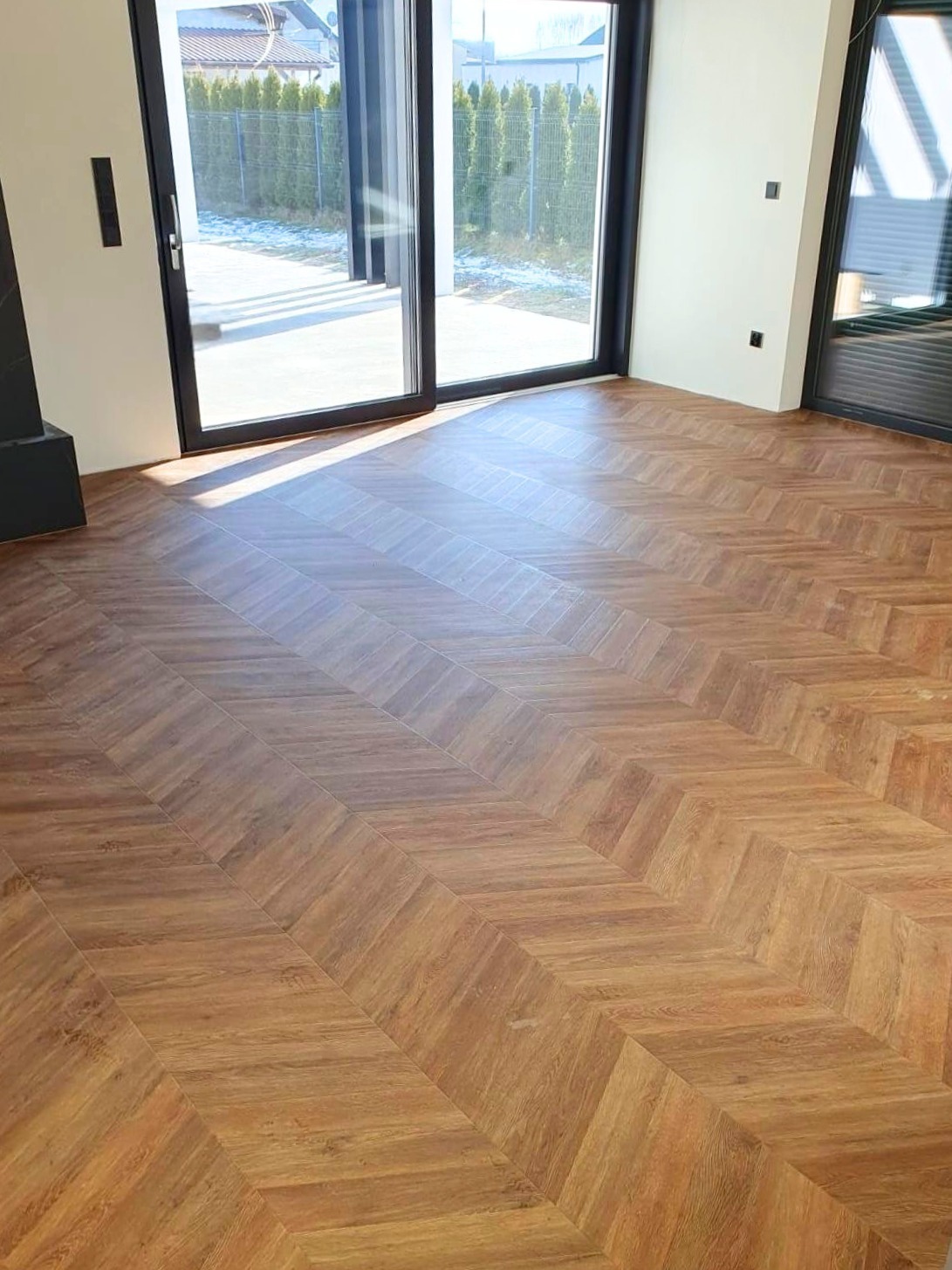 Nowoczesne wnętrze z podłogą ułożoną w jodełkę z paneli o ciepłym, brązowym odcieniu. Widoczne duże okna przesuwne z czarnymi ramami i minimalistyczne ściany.