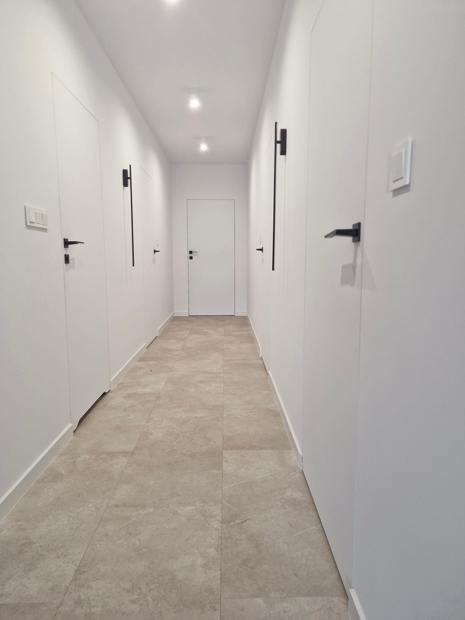 Nowoczesny korytarz z białymi, minimalistycznymi drzwiami i beżową podłogą z płytek. Czarne, proste klamki i oświetlenie punktowe.