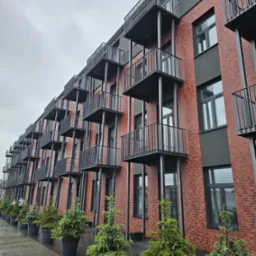 Nowoczesny budynek mieszkalny z czerwonej cegły z czarnymi balkonami i oknami, udekorowany rzędem małych drzewek iglastych w donicach wzdłuż chodnika.