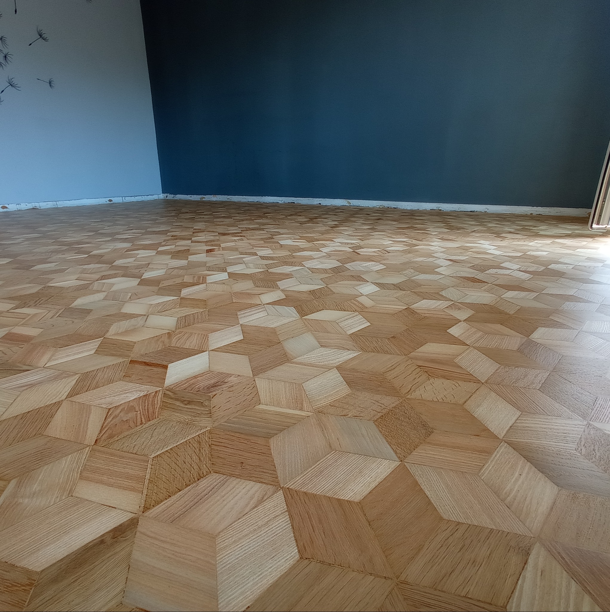 Ułożony parkiet z efektem 3D, jasne drewno, widok z perspektywy podłogi, szara ściana z motywem dmuchawca w tle.