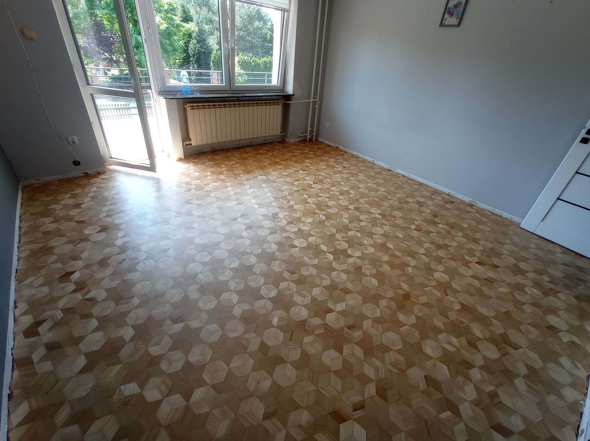 Wnętrze pokoju z ułożonym parkietem w geometryczny wzór kostki sześciennej; widoczne okno balkonowe, grzejnik i fragment drzwi.