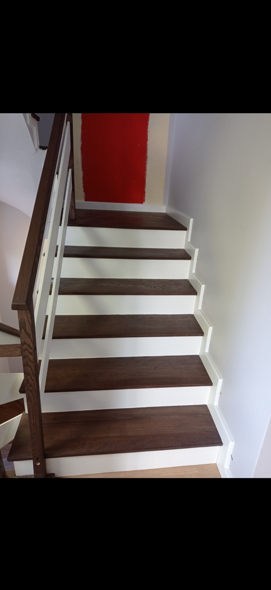 Nowoczesne schody drewniane z ciemnymi stopniami i białymi podstopnicami, widoczne fragmenty pomalowanej na czerwono ściany w tle.