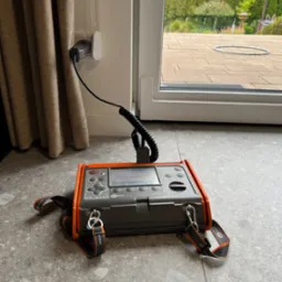 Miernik elektryczny Sonel MPI-520 podłączony do gniazdka elektrycznego, widoczny ekran z odczytem napięcia, tło stanowią zasłony i widok na ogród przez okno.