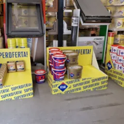 Ekspozycja w sklepie budowlanym z różnymi produktami marki Atlas: zaprawy, kleje, tynki w wiadrach i kanistrach, na żółtych standach z napisem 'Super Oferta!'