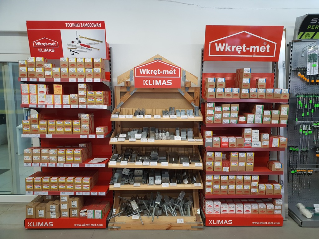 Ekspozycja w sklepie z technikami zamocowań Wkręt-met Klimas, prezentująca różnorodne elementy złączne na czerwonych półkach, z logo firmy widocznym na górze i dole ekspozycji.