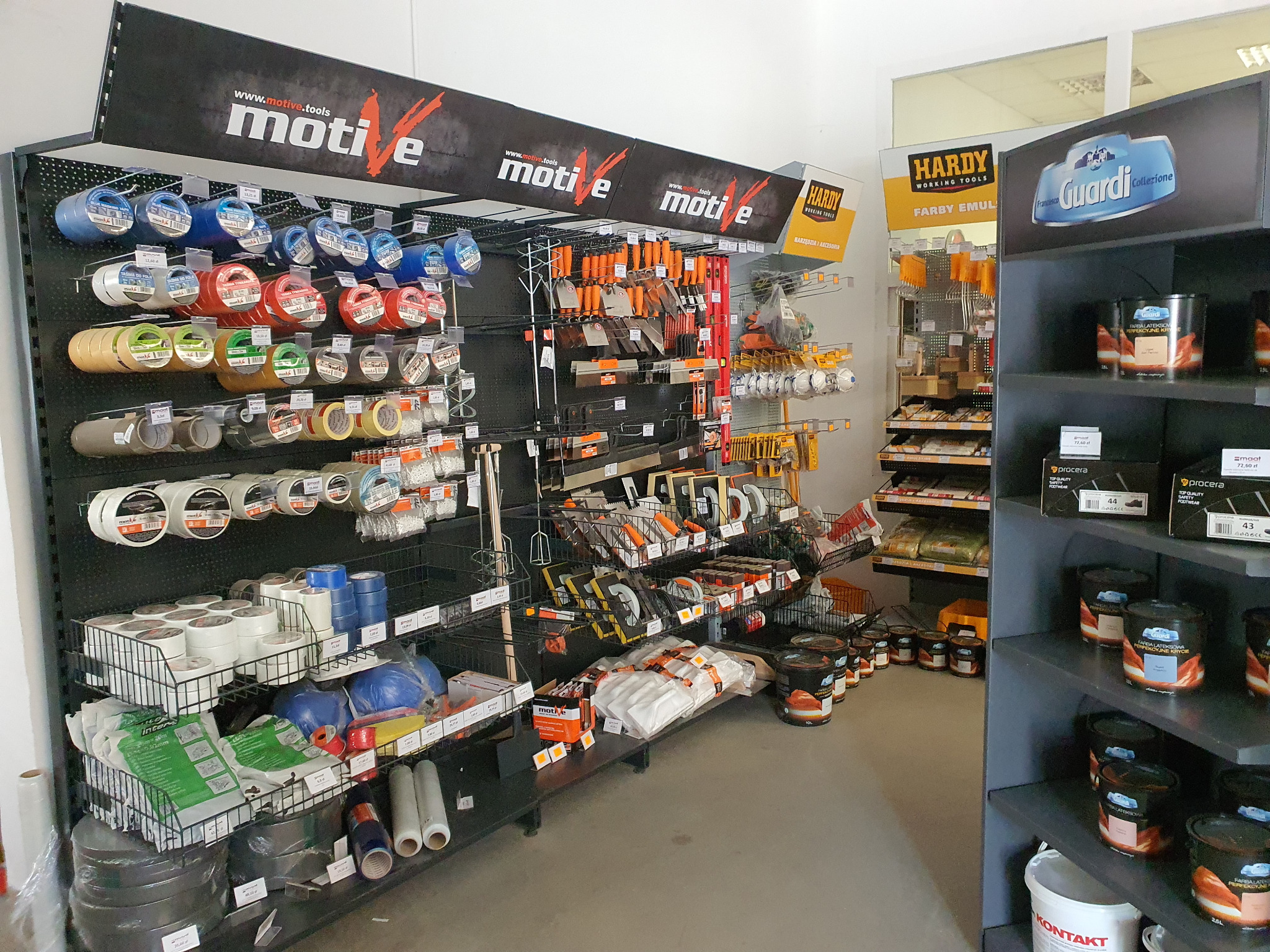 Wnętrze sklepu z materiałami budowlanymi, widoczne regały z taśmami malarskimi, narzędziami malarskimi, farbami marki Guardi i Hardy, oraz akcesoriami budowlanymi pod szyldem Motive.