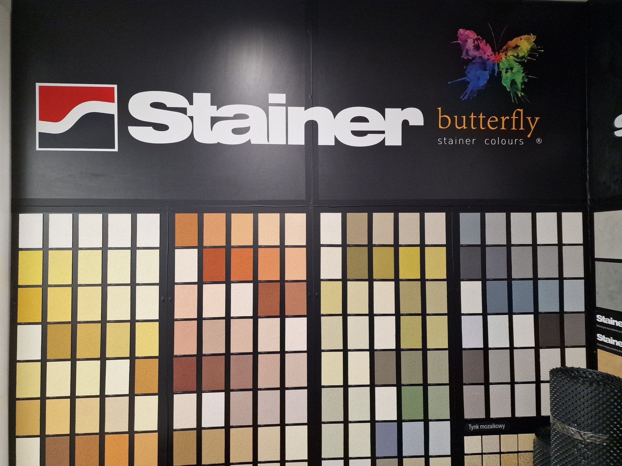 Ekspozycja próbek tynków dekoracyjnych Stainer Butterfly z logo firmy przedstawiającym kolorowego motyla.