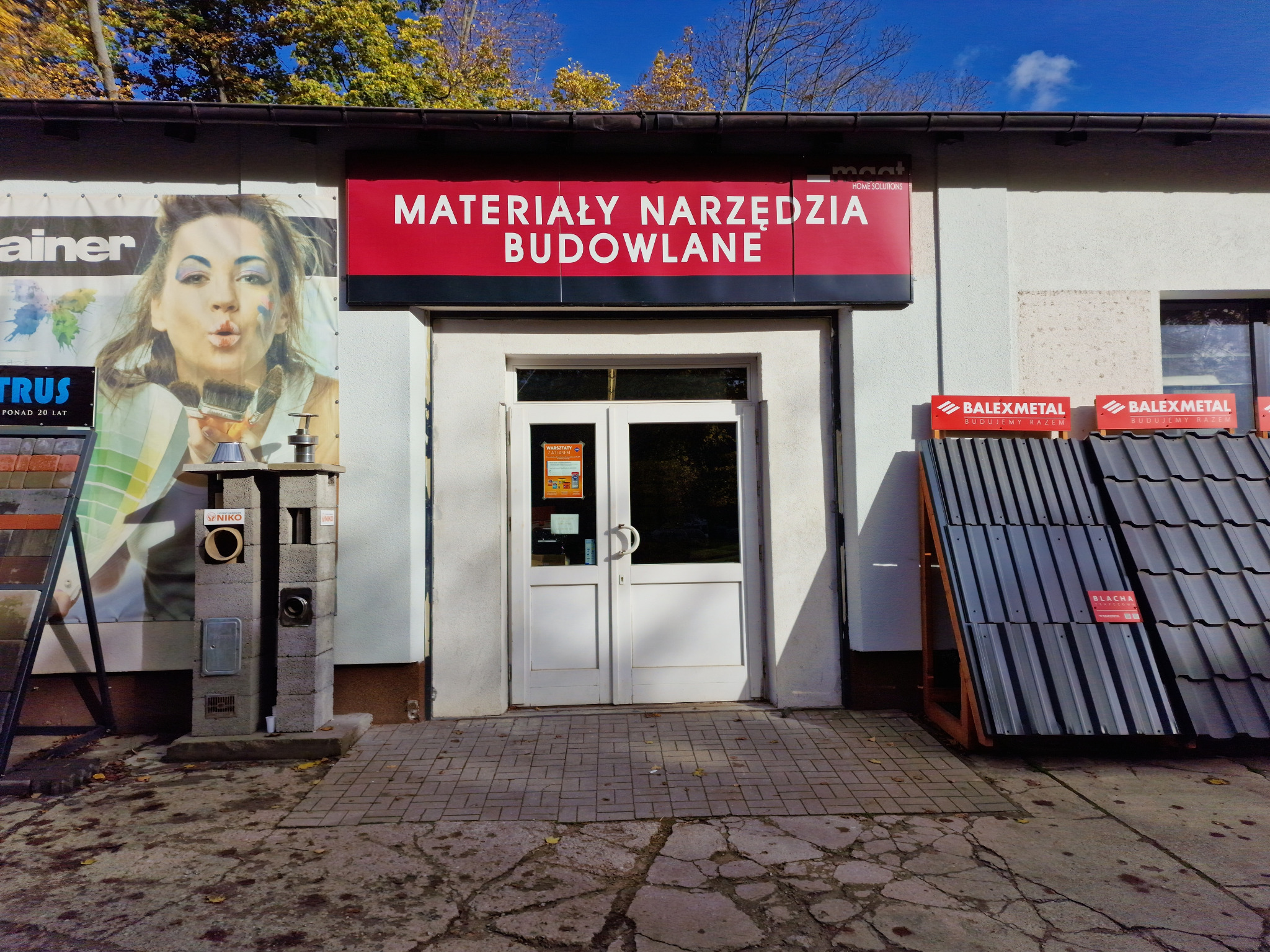 Fasada budynku z szyldem 'Materiały Narzędzia Budowlane', ekspozycja dachówki i blachy trapezowej oraz reklama z wizerunkiem kobiety i paletą kolorów, jesienne drzewa w tle.