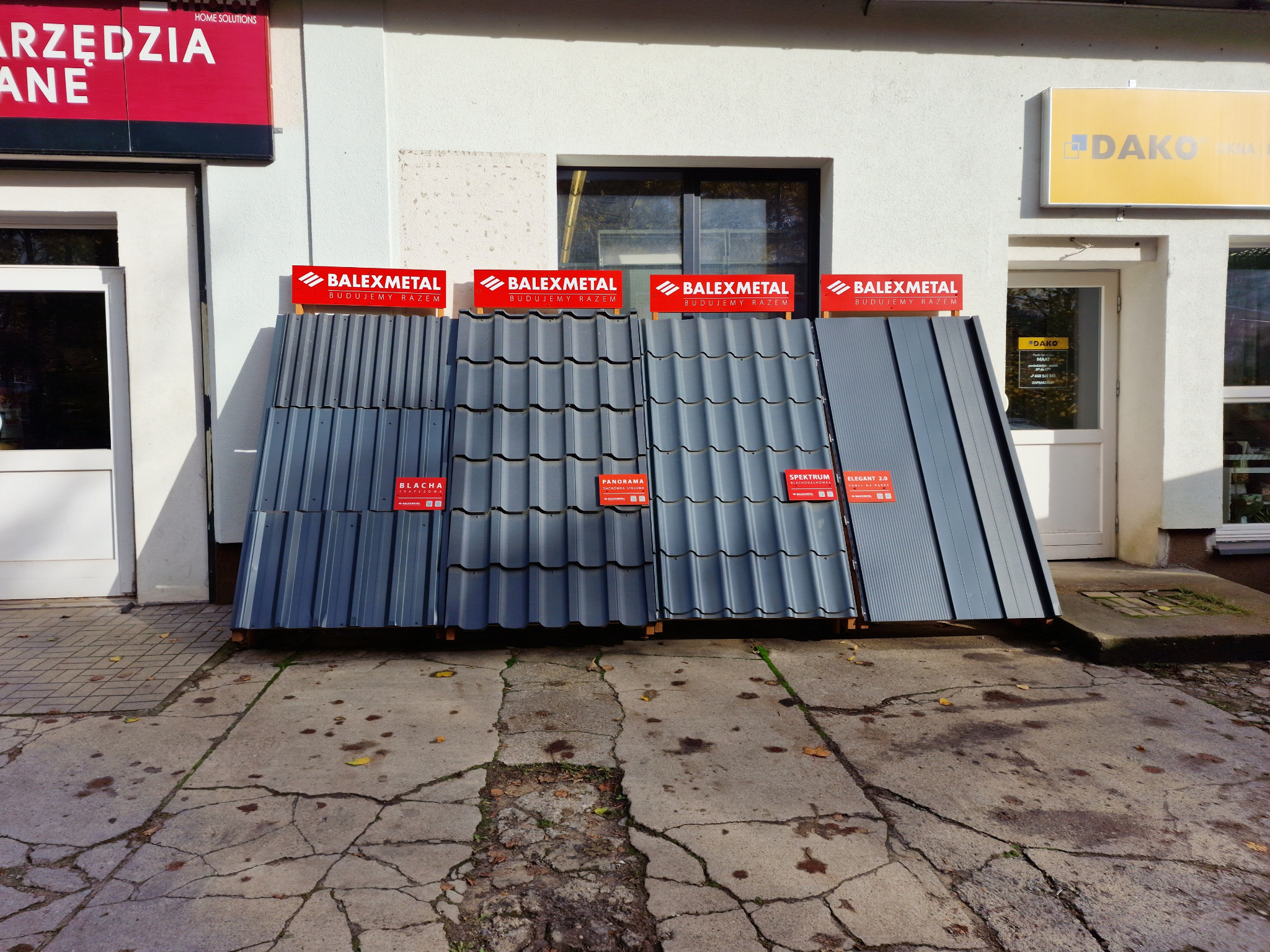 Ekspozycja różnych modeli metalowych pokryć dachowych marki BalexMetal przed budynkiem, prezentująca blachę trapezową i dachówkę metalową w odcieniach szarości.