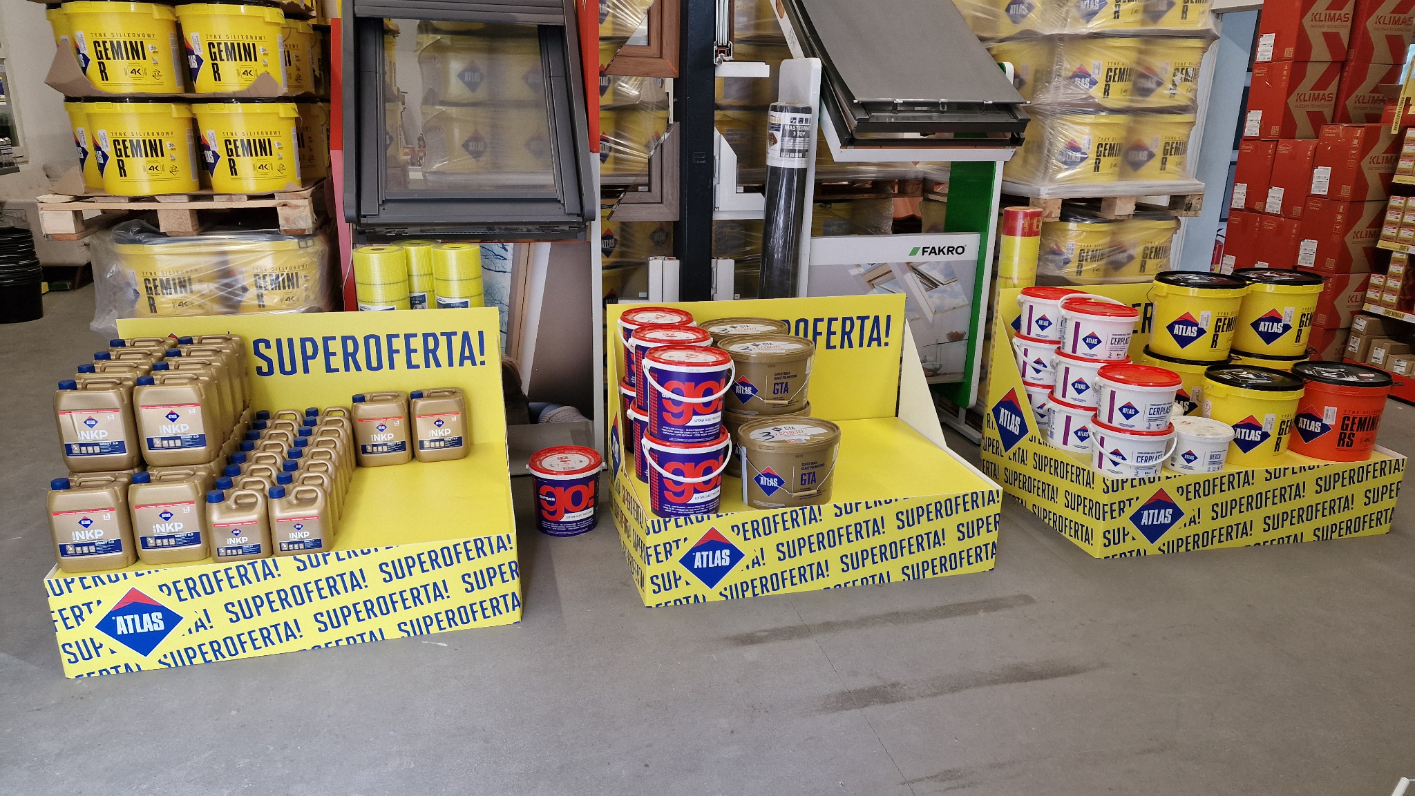 Ekspozycja w sklepie budowlanym z różnymi produktami marki Atlas: zaprawy, kleje, tynki w wiadrach i kanistrach, na żółtych standach z napisem 'Super Oferta!'