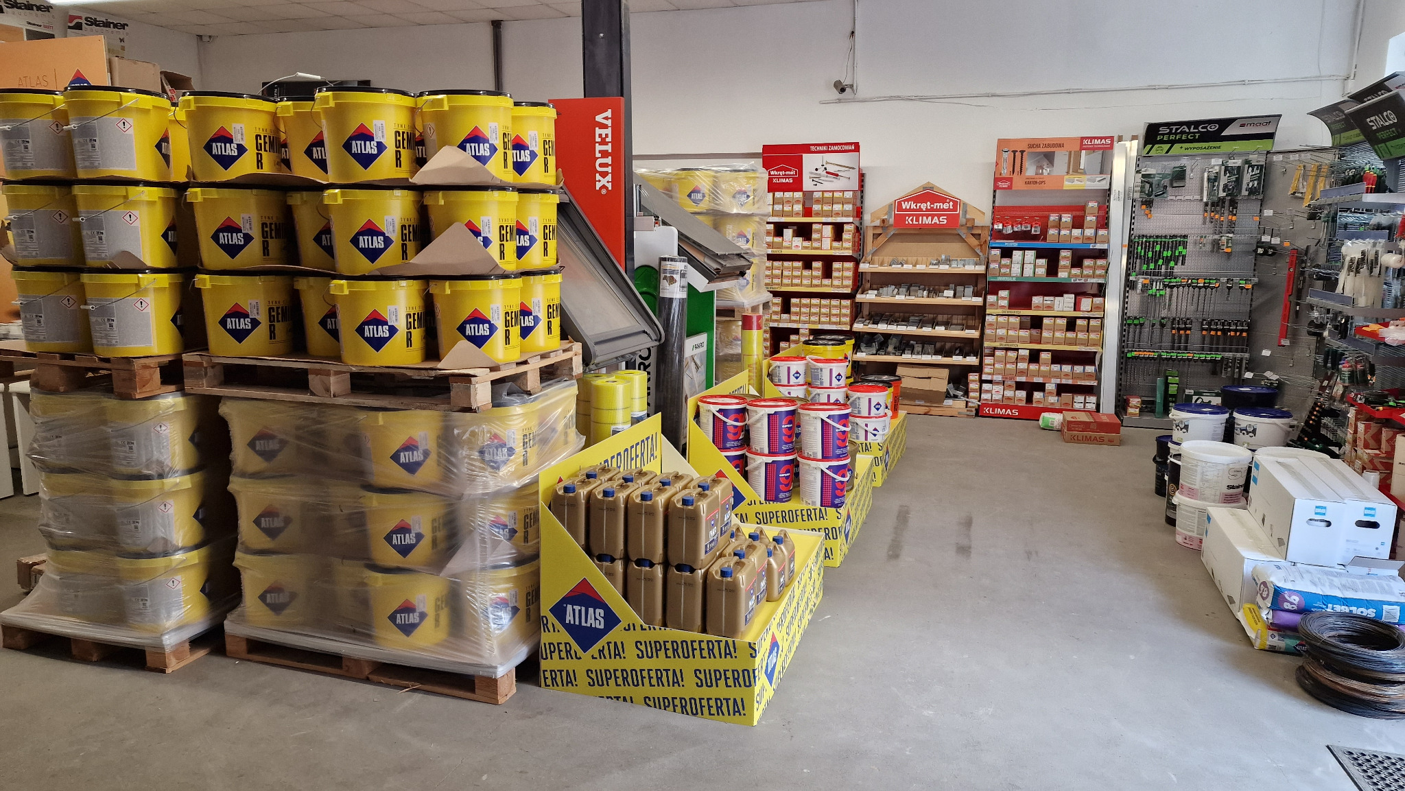 Wnętrze składu budowlanego w Gorzowie Wielkopolskim, z paletami żółtych wiader z klejem Atlas i brązowych kanistrów, regały z narzędziami i akcesoriami budowlanymi w tle.