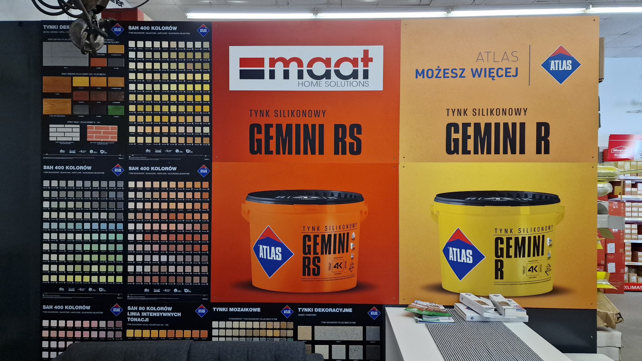 Ekspozycja reklamowa tynków silikonowych Atlas Gemini RS i Gemini R na stoisku w sklepie budowlanym, z paletami kolorów i logo Maat Home Solutions.