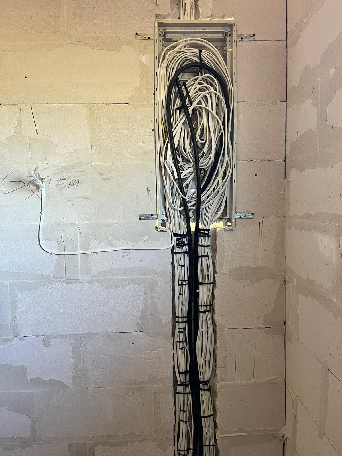 Pionowy kanał w ścianie z betonu komórkowego wypełniony wiązką białych i czarnych przewodów elektrycznych, zabezpieczonych czarnymi opaskami zaciskowymi.