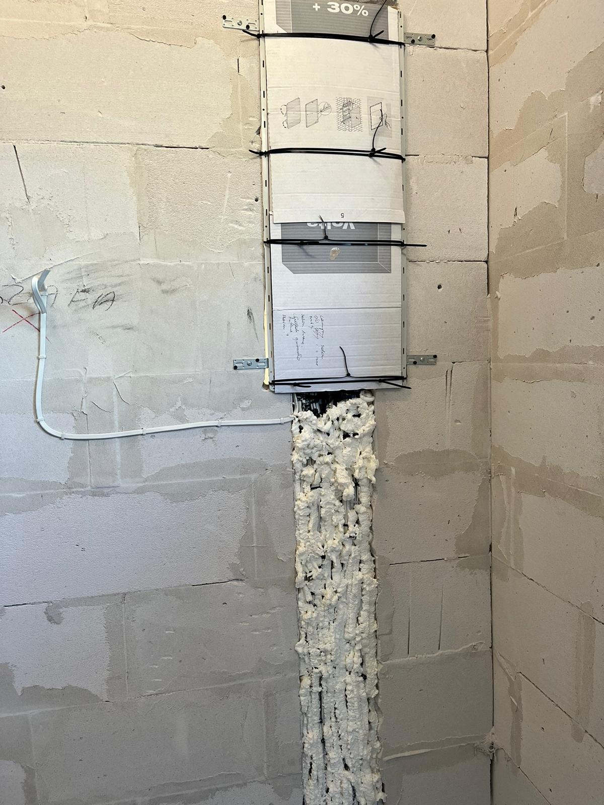 Pionowy wykop w murze z szarego betonu wypełniony pianką montażową, zabezpieczony kartonem z instrukcjami i notatkami, przymocowanym czarnymi opaskami zaciskowymi do metalowych wsporników.