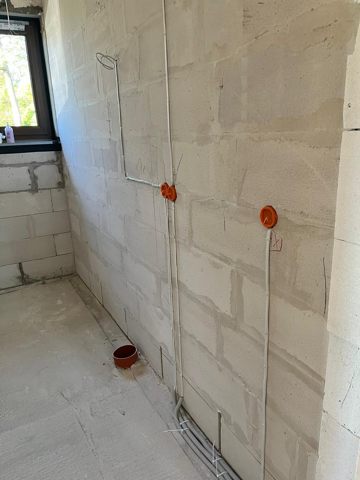 Surowy stan ścian z betonu komórkowego z widoczną instalacją elektryczną w postaci białych kabli i pomarańczowych puszek pod gniazdka, częściowo poprowadzonych w podłodze w rurach karbowanych.