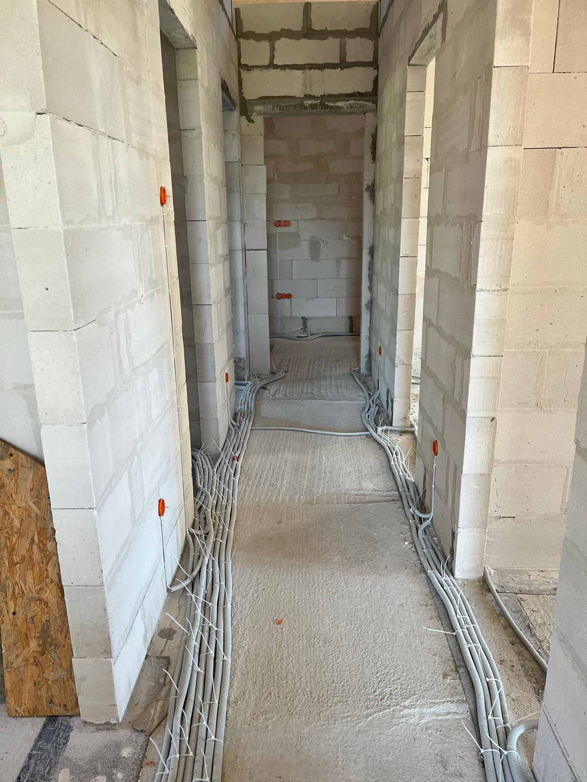 Wnętrze w stanie surowym z widocznymi przewodami elektrycznymi ułożonymi w korytach wzdłuż ścian, pomarańczowe puszki instalacyjne w ścianach z betonu komórkowego.