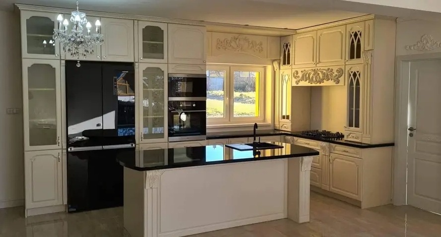Elegancka kuchnia w odcieniach kremu z czarnym blatem i wyspą kuchenną, zdobiona sztukaterią i oświetlona kryształowym żyrandolem, z widocznym oknem i zabudowanym sprzętem AGD.