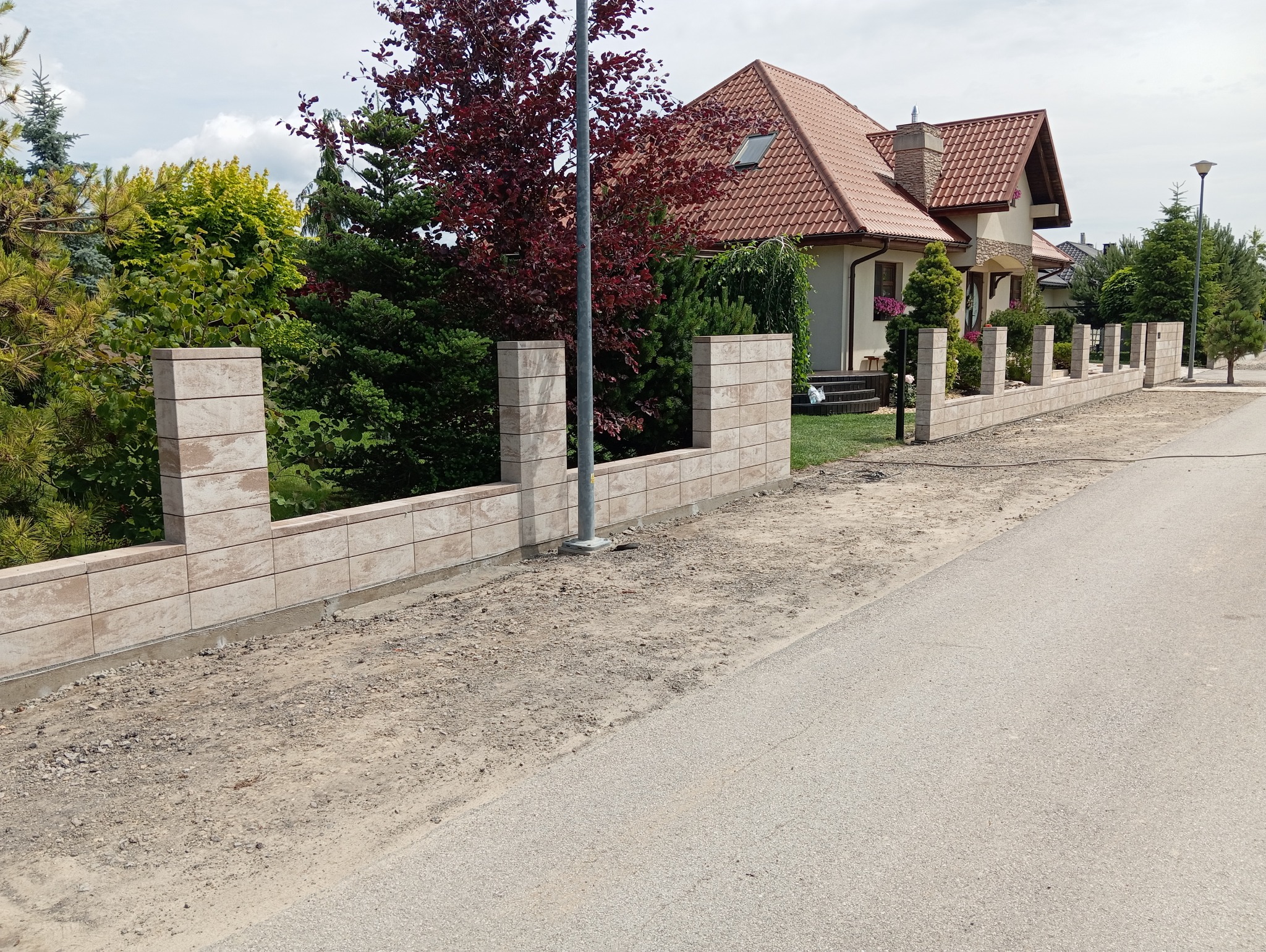 Nowoczesne ogrodzenie z paneli betonowych imitujących kamień, oddzielające posesję od drogi, widoczne ślady prac ziemnych i przygotowania terenu.