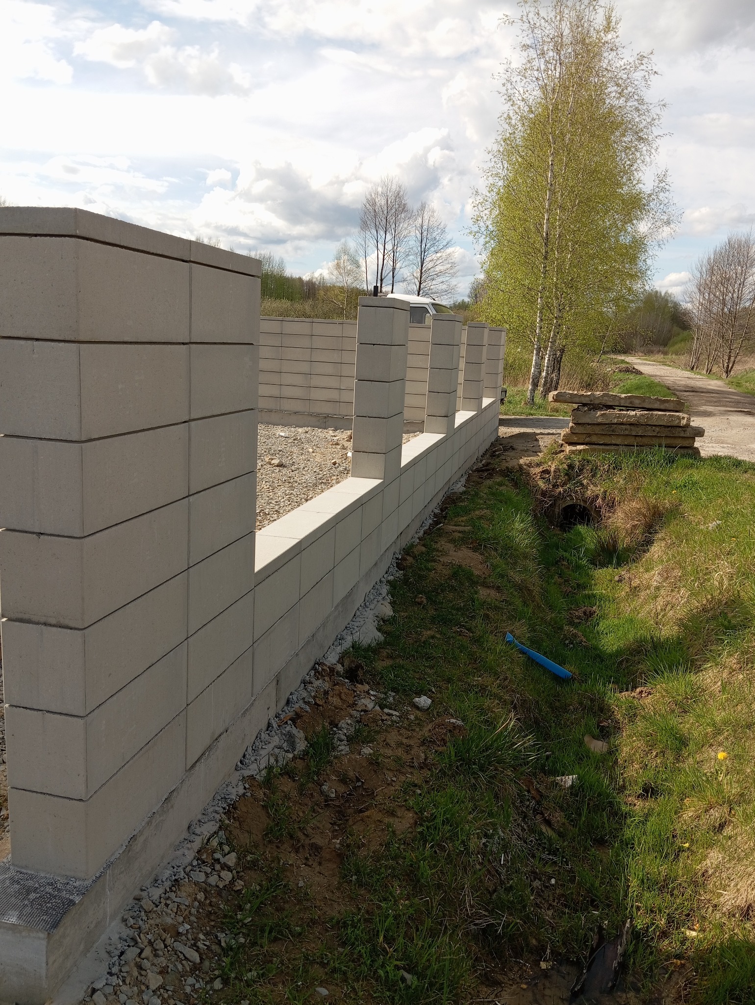 Wznoszone ogrodzenie modułowe z szarego betonu, widoczny fundament, teren przygotowany pod zasypanie żwirem, w tle brzozy i pochmurne niebo.