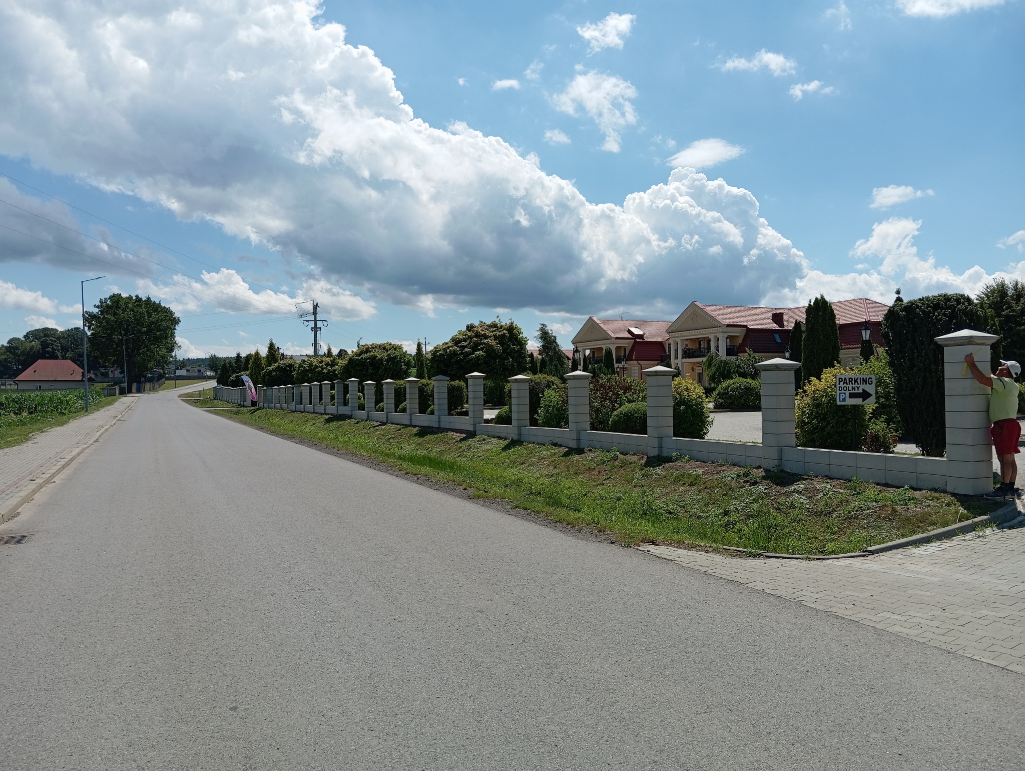 Długi, niski, betonowy mur z kolumnami oddzielający posesję z budynkiem w tle od drogi, pracownik poprawia jego wykończenie młotkiem.