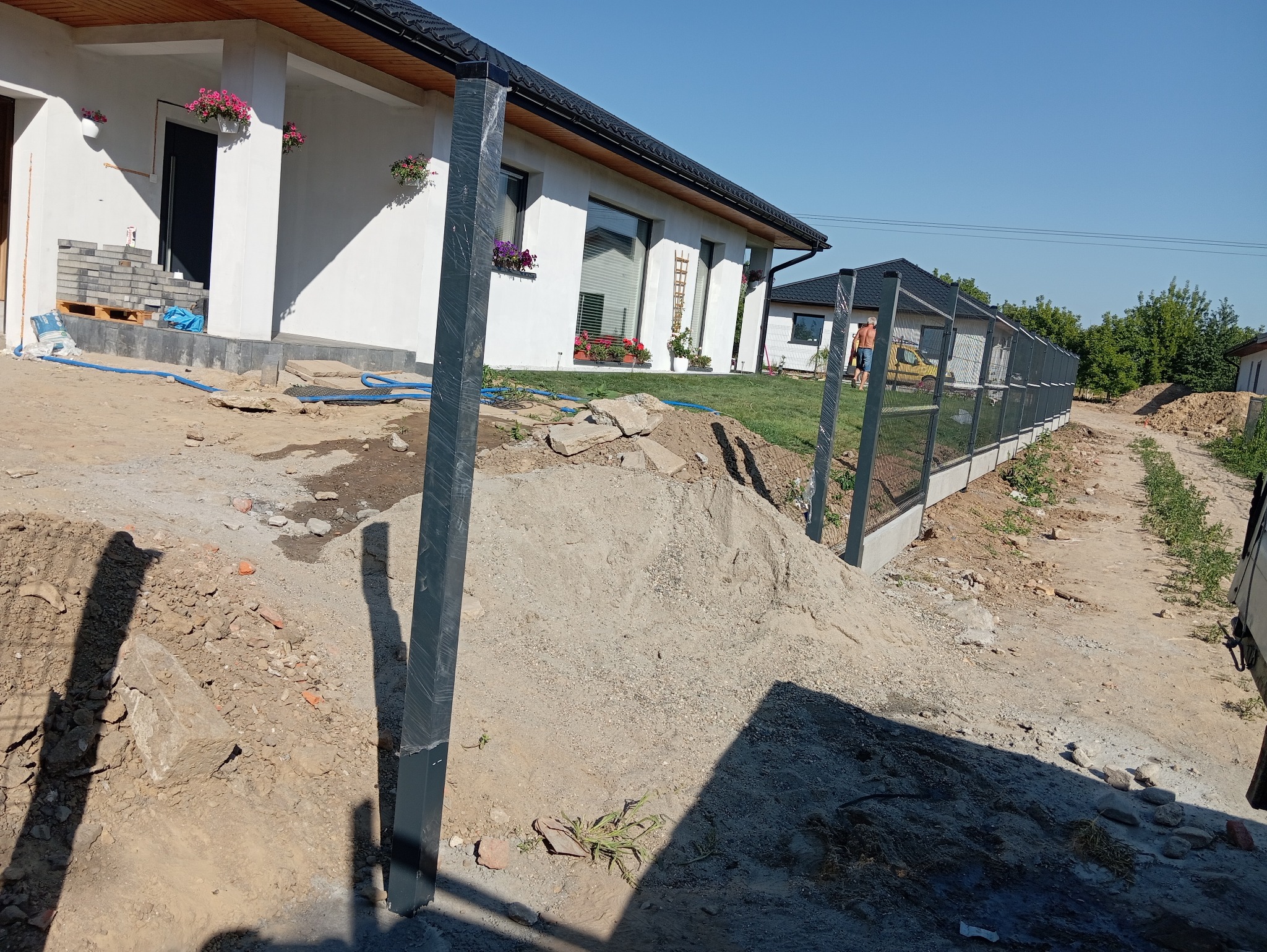 W trakcie montażu ogrodzenia panelowego: widoczne słupki w folii ochronnej, częściowo zamontowane panele na betonowej podmurówce, przygotowany teren z hałdą piasku i gruzu przed nowym domem.