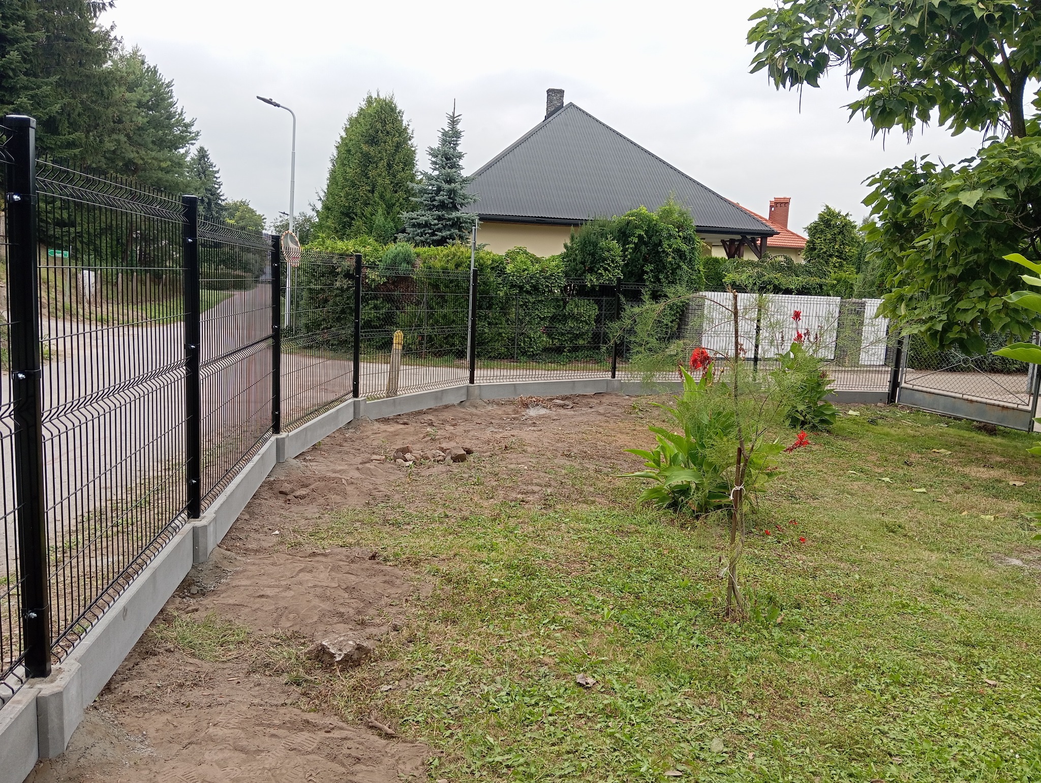 Nowoczesne ogrodzenie panelowe z podmurówką betonową, widok na posesję z posadzoną roślinnością i bramą w tle.