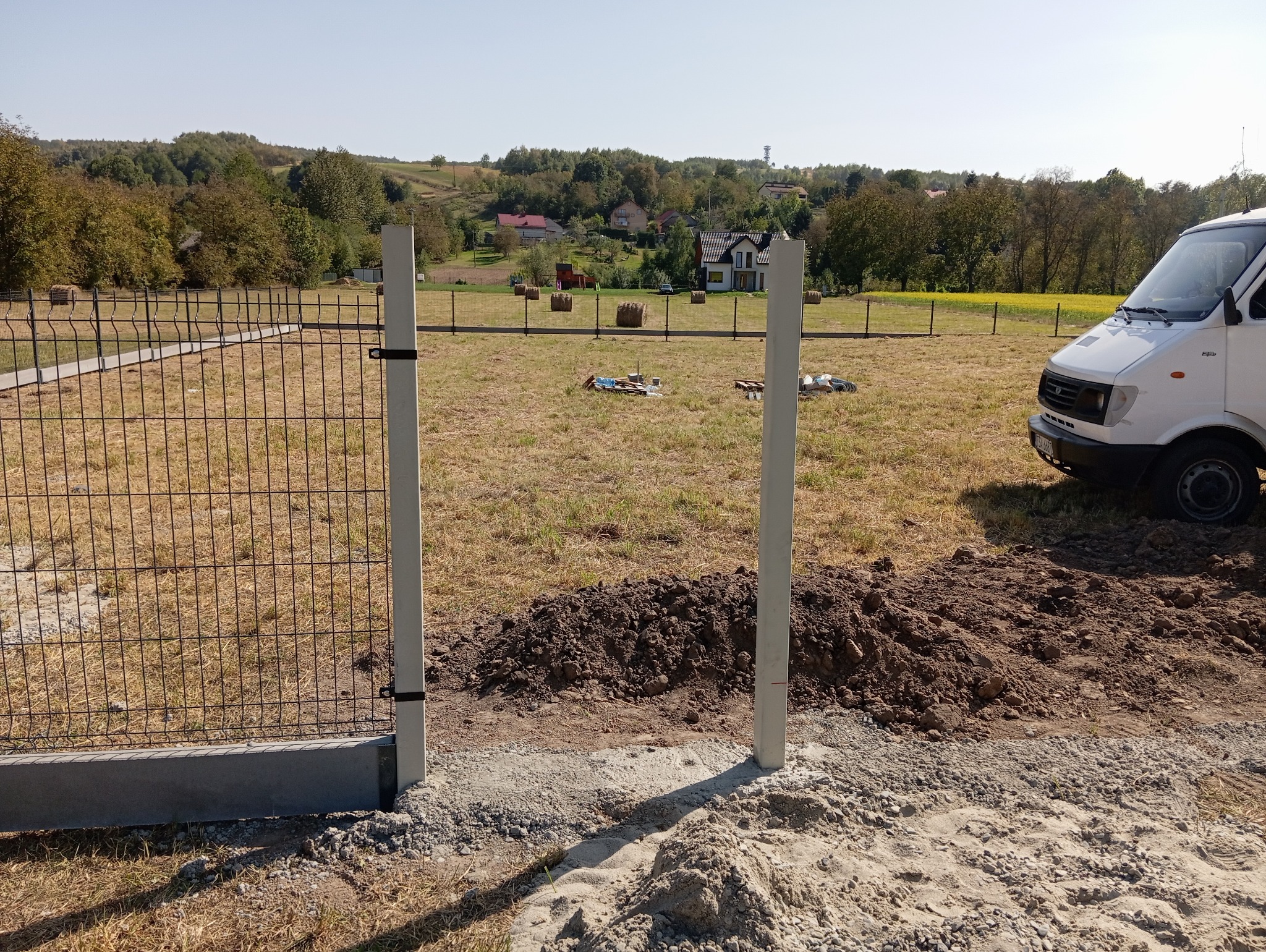 Trwa montaż ogrodzenia panelowego: widoczne słupki i fragment zamontowanego panelu na tle wiejskiego krajobrazu z białym busem parkującym obok.