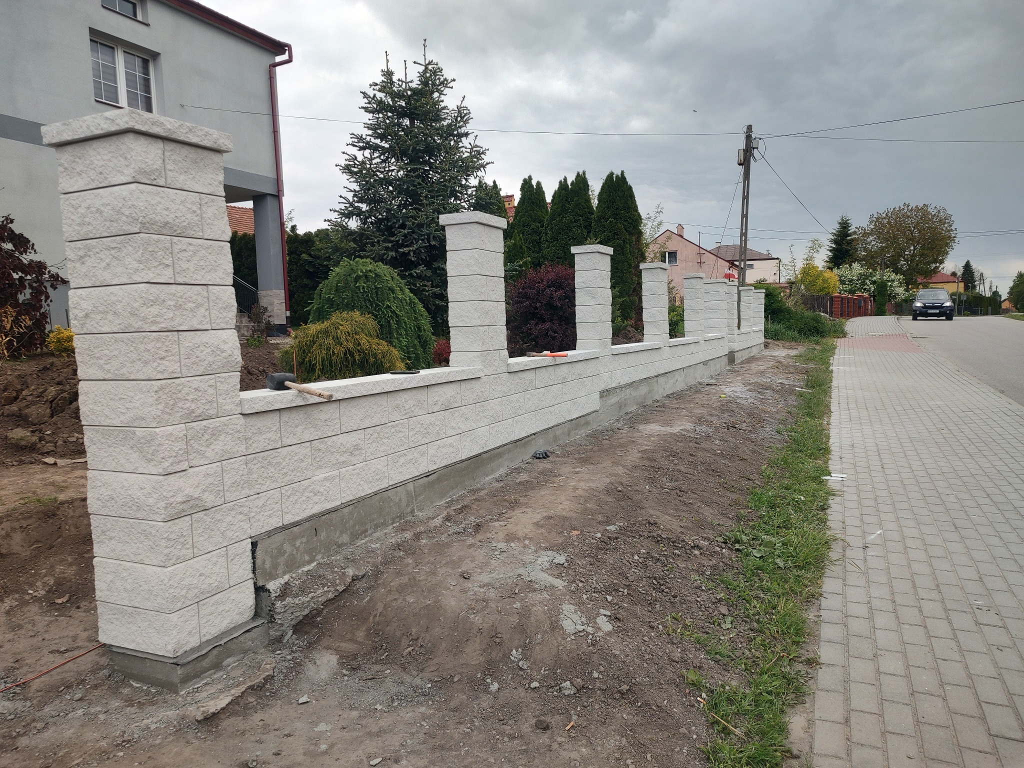 Nowoczesne ogrodzenie modułowe z jasnych bloczków betonowych, z widocznymi słupkami i podmurówką, oddzielające posesję od chodnika. Tło stanowią drzewa i zabudowa jednorodzinna.