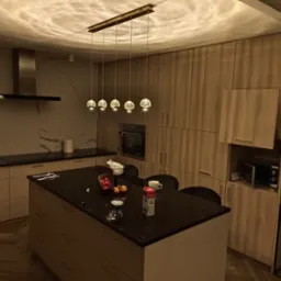 Nowoczesna kuchnia z wyspą, oświetlona wiszącymi lampami o kulistych kloszach, z widocznymi elementami wyposażenia, takimi jak okap, piekarnik i mikrofalówka, oraz akcentami dekoracyjnymi, w tym...