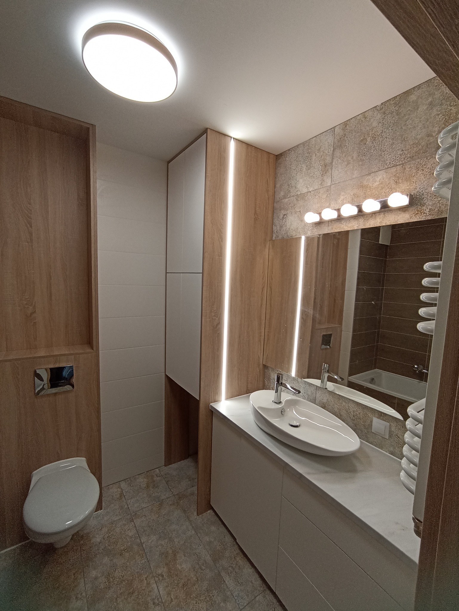 Nowoczesna łazienka z owalną umywalką nablatową, podświetlanym lustrem i drewnianymi akcentami, widok na wannę w odbiciu lustra.