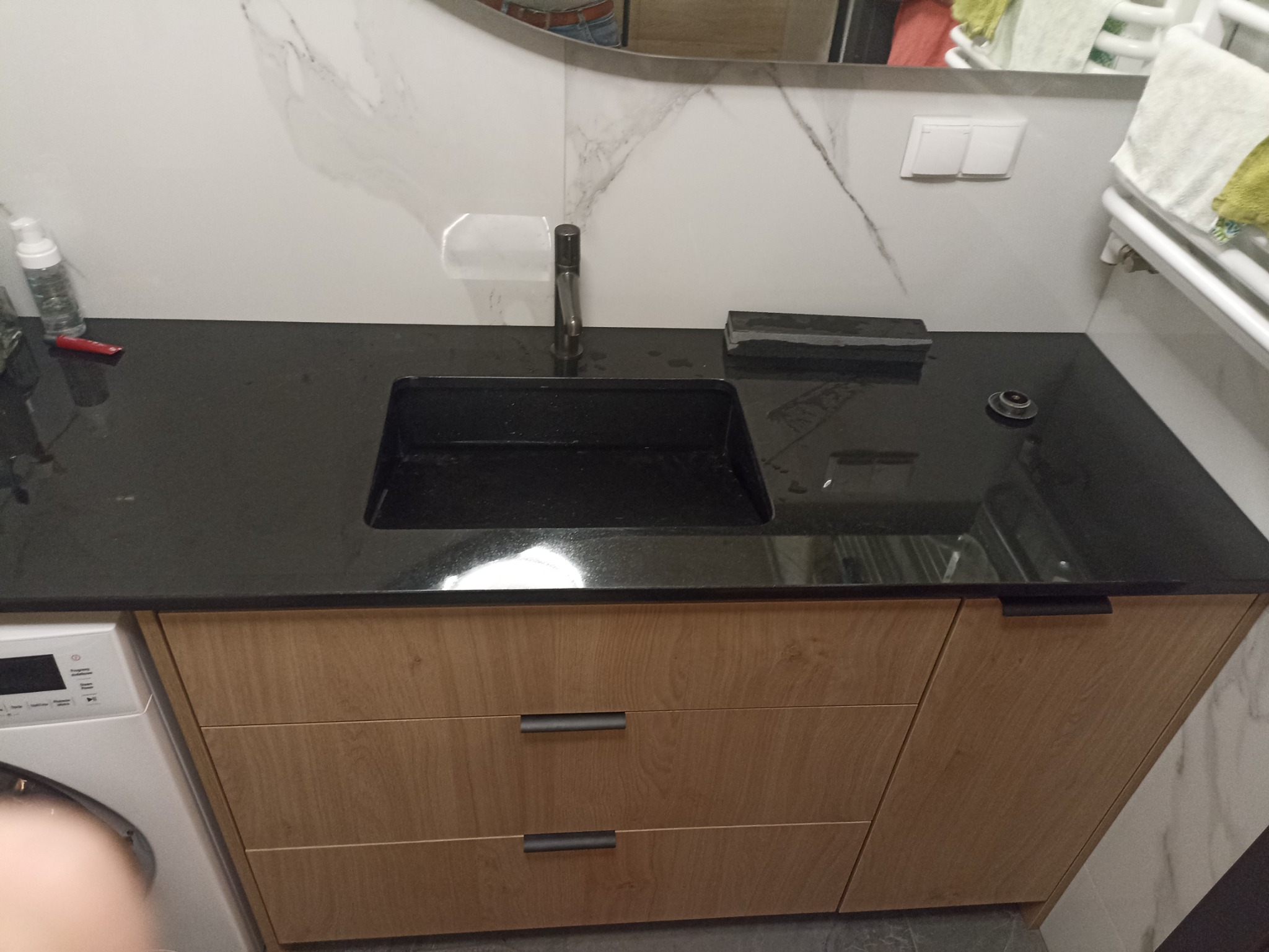 Czarna, kamienna umywalka wpuszczana w drewnianą szafkę łazienkową z trzema szufladami, widoczna bateria, dozownik mydła i fragment pralki.