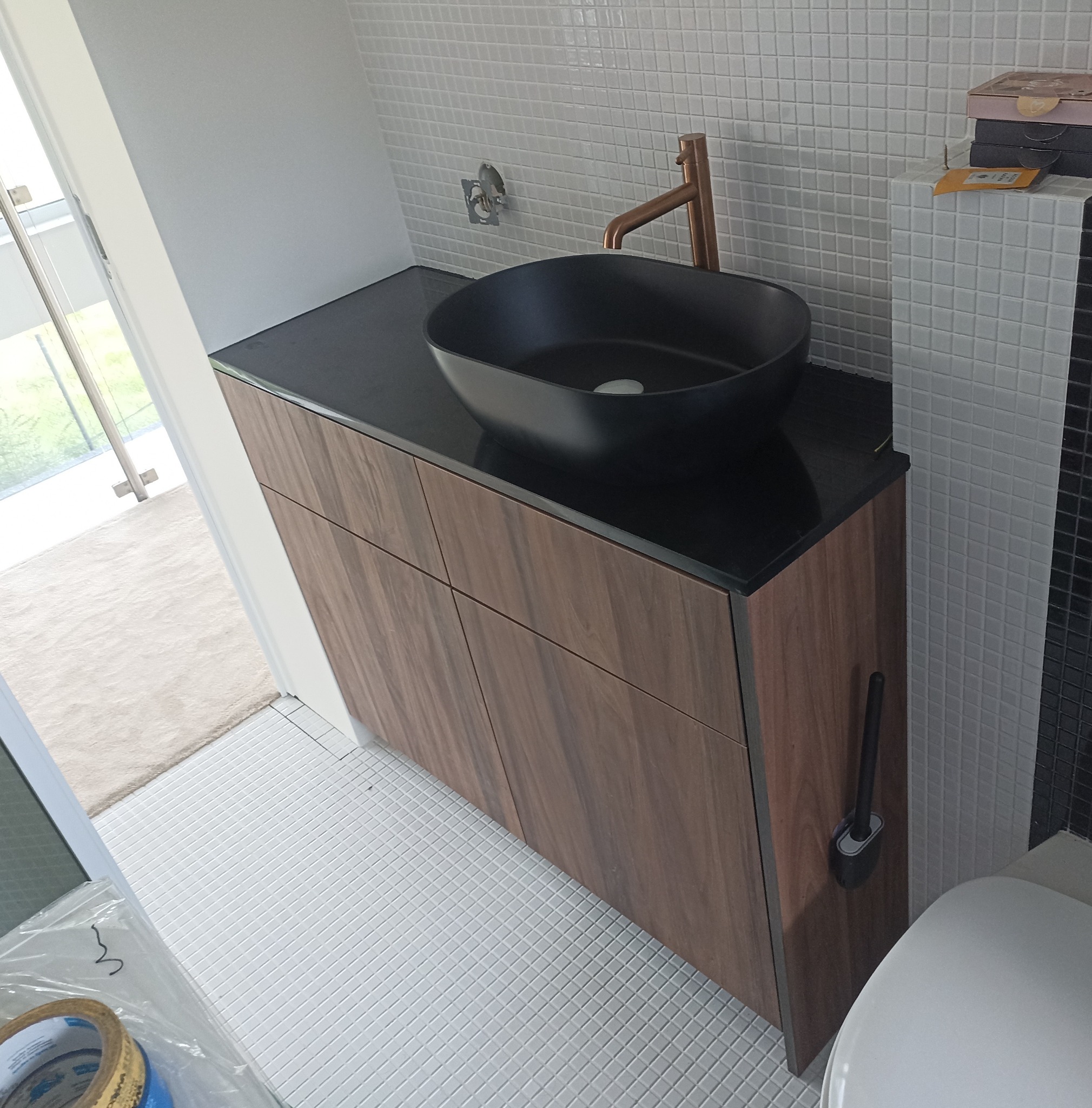 Nowoczesna łazienka po remoncie: czarna umywalka nablatowa z miedzianą baterią na blacie z ciemnego kamienia, szafka z drewnopodobnymi frontami, białe mozaikowe płytki na ścianach i podłodze...