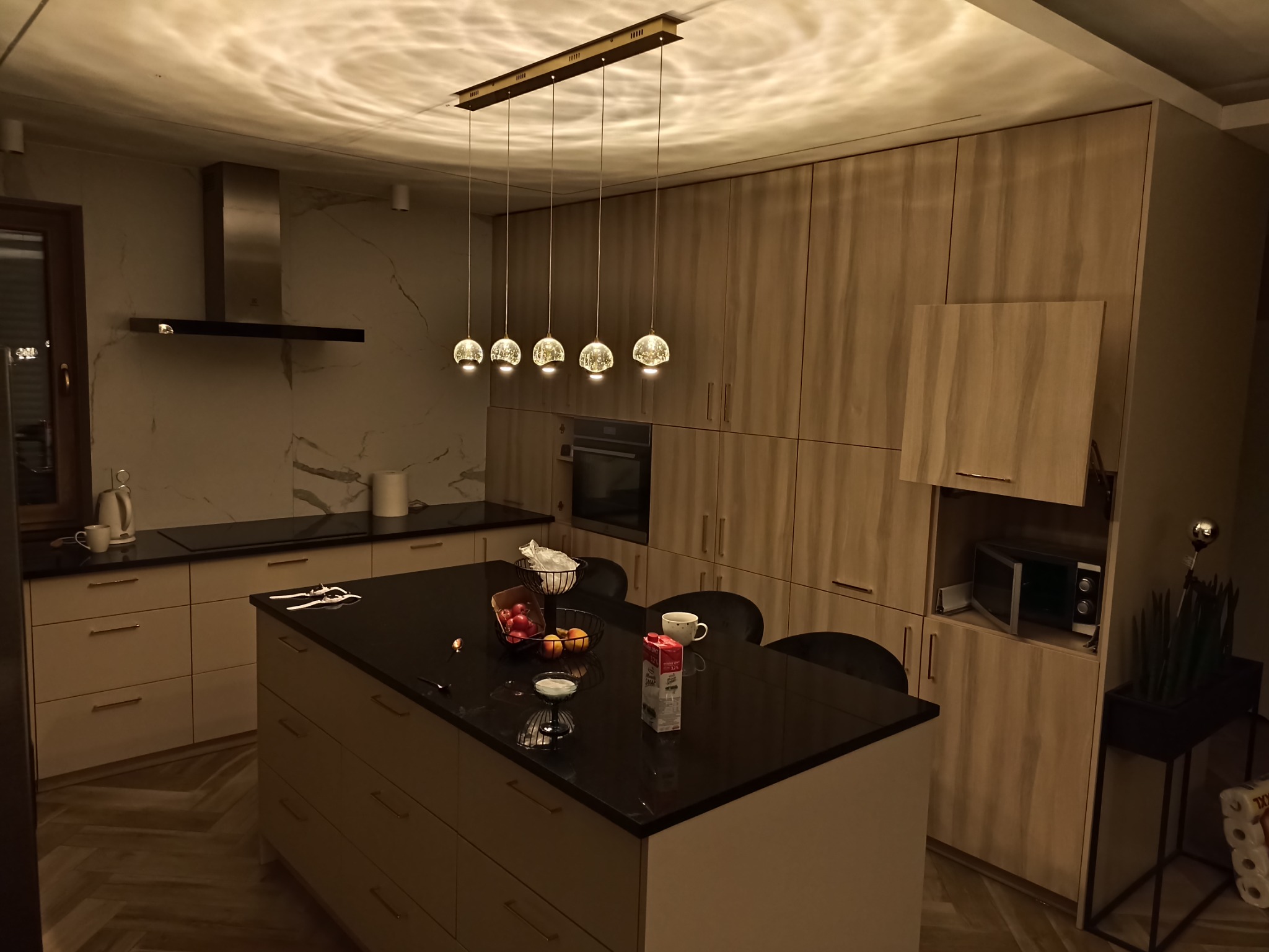 Nowoczesna kuchnia z wyspą, oświetlona wiszącymi lampami o kulistych kloszach, z widocznymi elementami wyposażenia, takimi jak okap, piekarnik i mikrofalówka, oraz akcentami dekoracyjnymi, w tym...