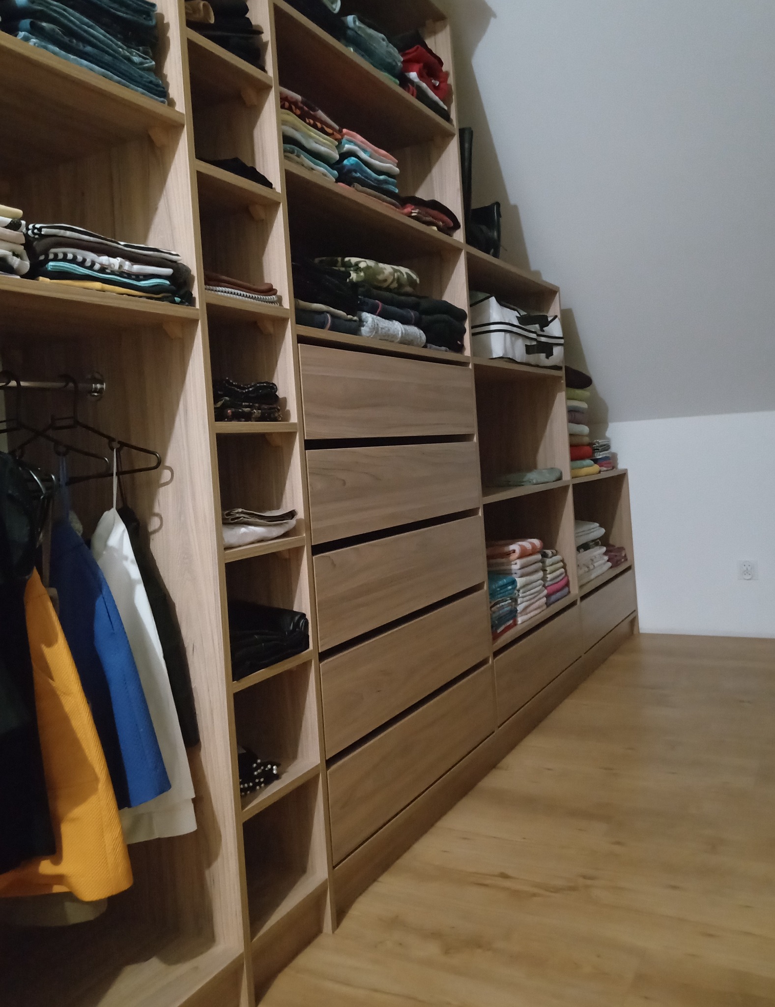 Wnętrze garderoby w skosie, z drewnianymi półkami i szufladami, wypełnione złożonymi ubraniami i wiszącymi koszulami na wieszakach.