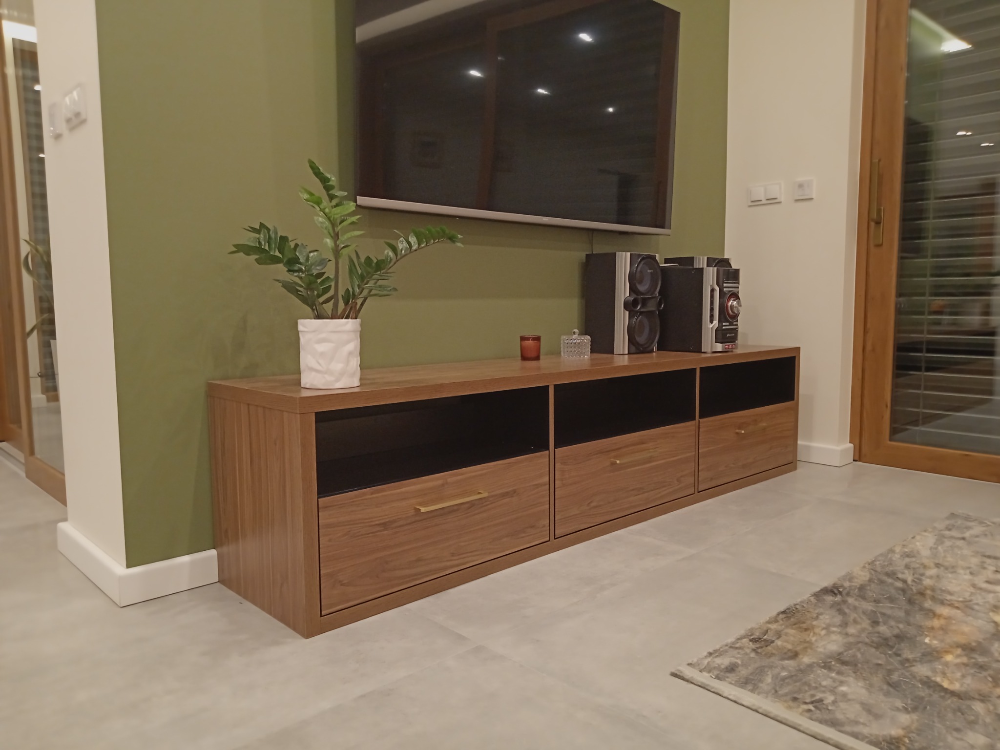 Nowoczesny salon z zieloną ścianą, drewnianą szafką RTV z szufladami, rośliną w białej doniczce i sprzętem audio.