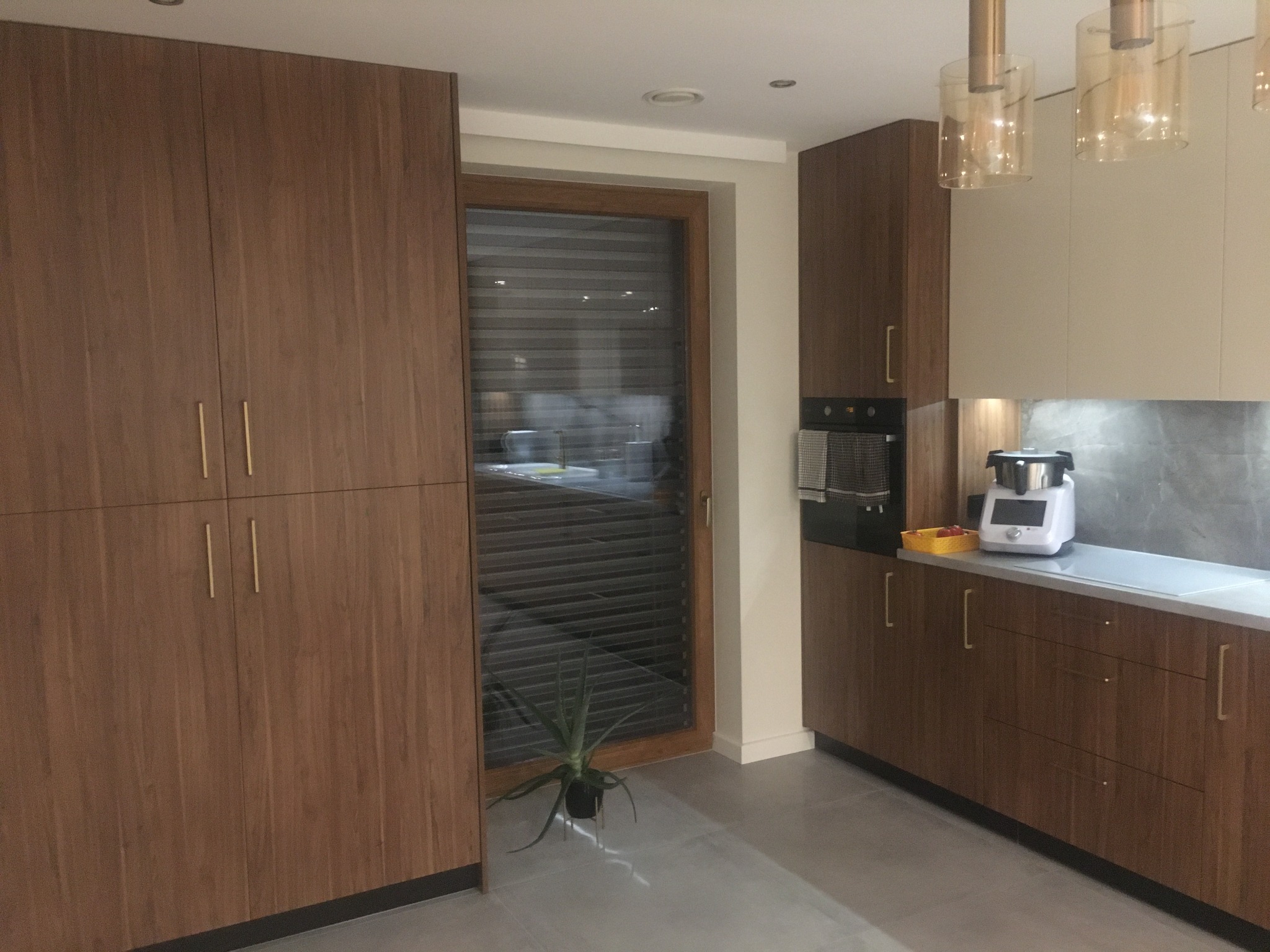 Nowoczesna kuchnia z drewnianymi szafkami i złotymi uchwytami, widok na okno z żaluzjami i roślinę w doniczce na podłodze.