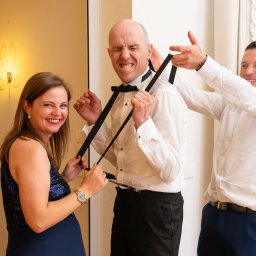 Machnicki Fotografia - Uśmiechnięta kobieta i mężczyzna pomagają mężczyźnie w białej koszuli założyć szelki i muchę, przygotowania do ślubu.