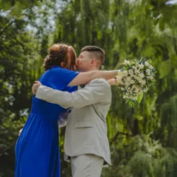 Para młoda w objęciach, panna młoda w kobaltowej sukience, pan młody w jasnym garniturze, bukiet z białych róż i niebieskich akcentów, pocałunek na tle zieleni drzew.