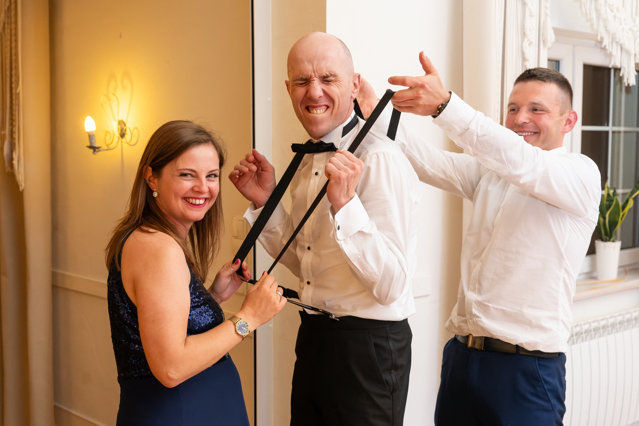 Uśmiechnięta kobieta i mężczyzna pomagają mężczyźnie w białej koszuli założyć szelki i muchę, przygotowania do ślubu.