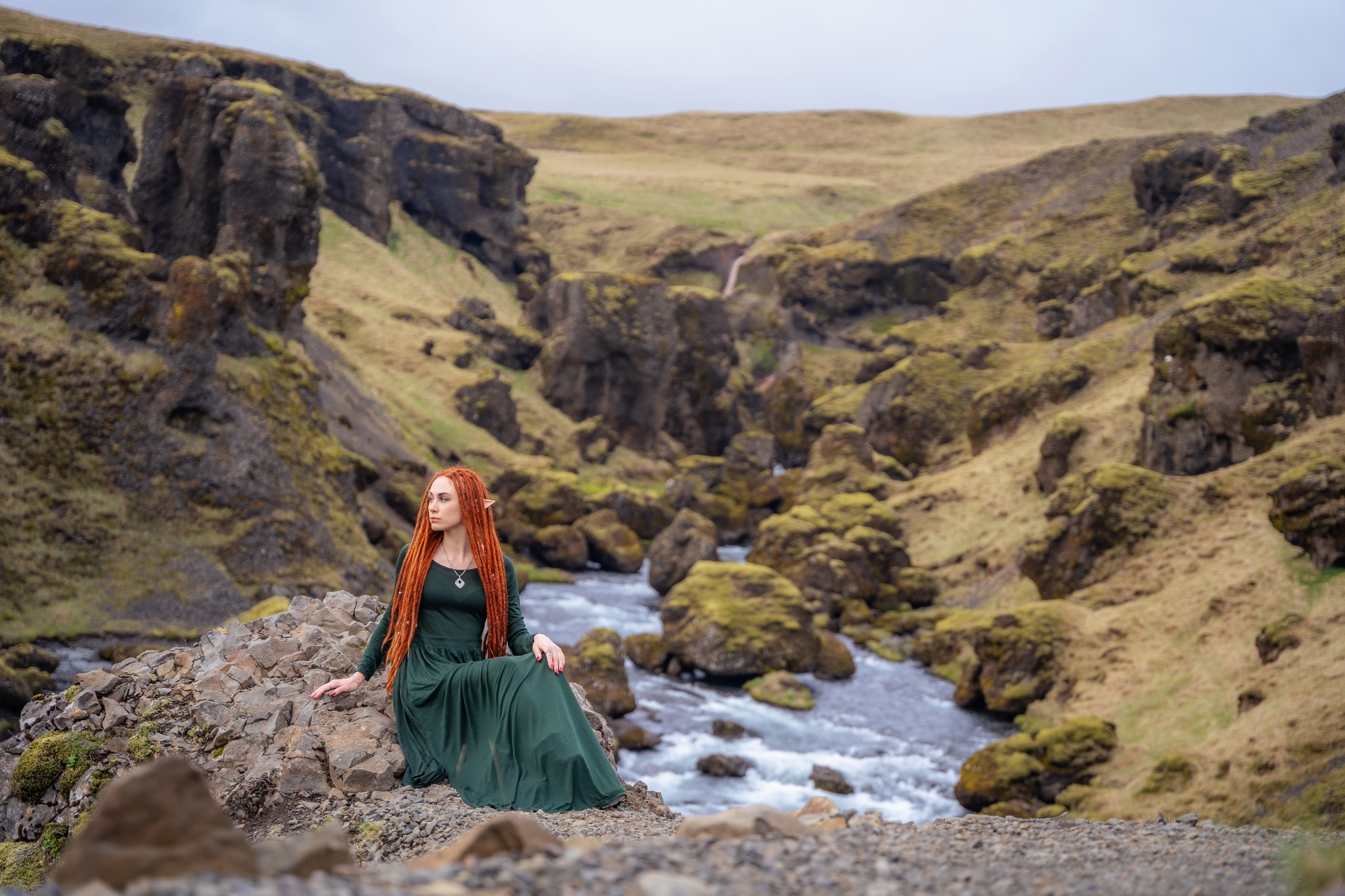 Kobieta z długimi, rudymi włosami i w zielonej sukni siedzi na kamieniach w islandzkim krajobrazie z rzeką i wzgórzami w tle.