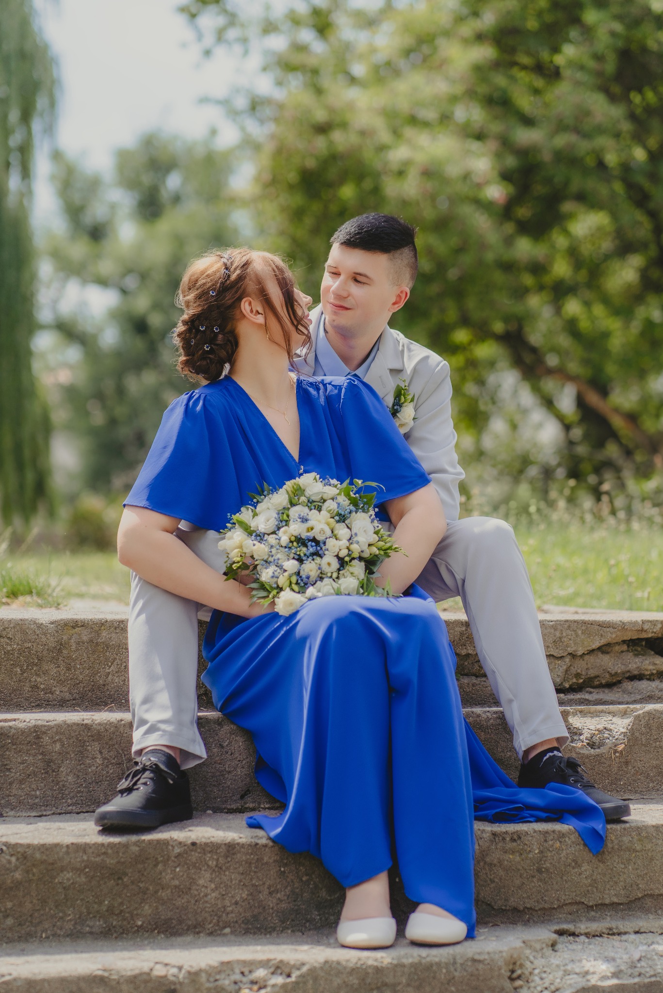 Para młoda siedząca na betonowych schodach w parku: panna młoda w kobaltowej sukni z bukietem białych i niebieskich kwiatów, pan młody w jasnoszarym garniturze, oboje patrzą na siebie z uśmiechem.