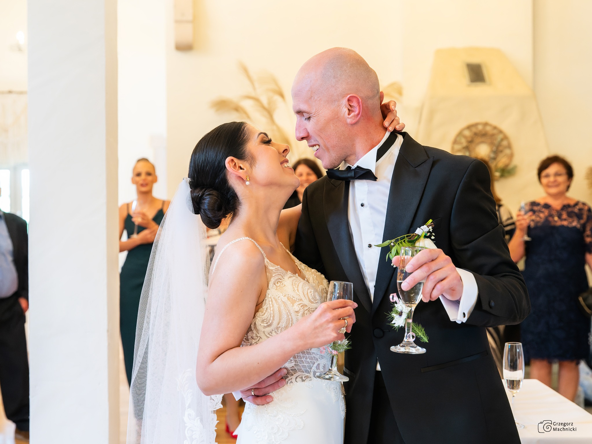 Uśmiechnięta para młoda w eleganckich strojach, trzymająca kieliszki z szampanem, patrzy na siebie czule w jasnym wnętrzu sali weselnej, w tle goście.