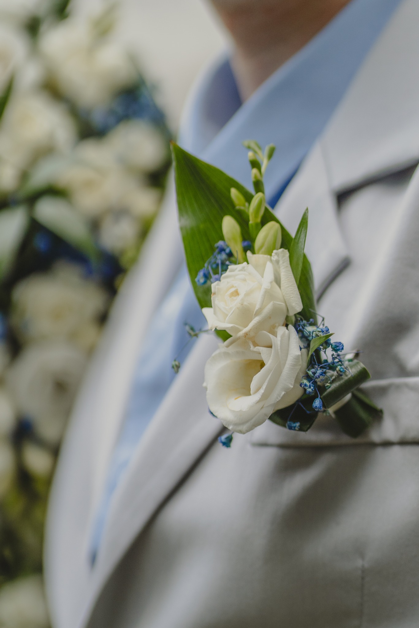 Biały butonierka z róż i niebieskich kwiatów na szarym garniturze z błękitną koszulą, z bukietem w tle.