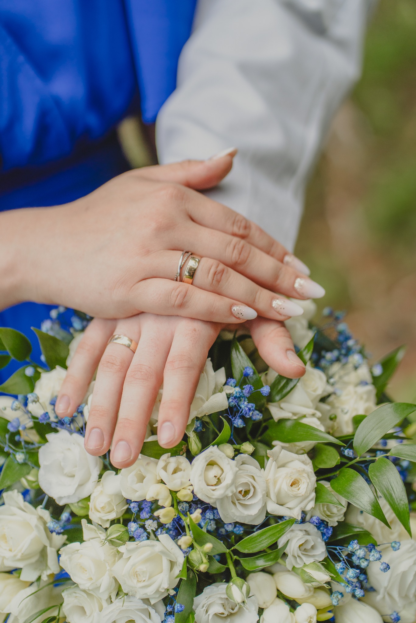 Dłonie pary młodej z obrączkami spoczywają na bukiecie ślubnym z białych róż i drobnych niebieskich kwiatów, manicure panny młodej z delikatnymi zdobieniami.