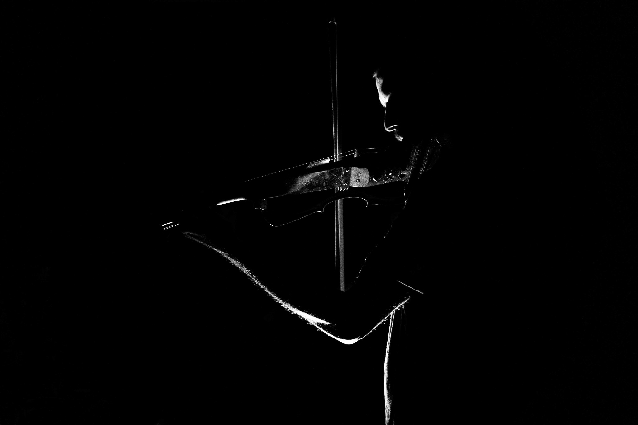 Silueta muzyka grającego na elektrycznych skrzypcach w ciemnym otoczeniu, z widocznym fragmentem twarzy i oświetlonymi konturami instrumentu.